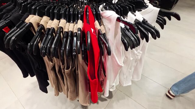 Person hand reaching for clothes on rack in clothing store with various colored tank tops including red white beige and black garments for sale display.