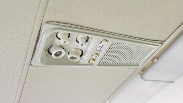 Commercial aircraft cabin overhead passenger service unit close up showing adjustable reading lights, individual air vents, and attendant call button on grey ceiling panel