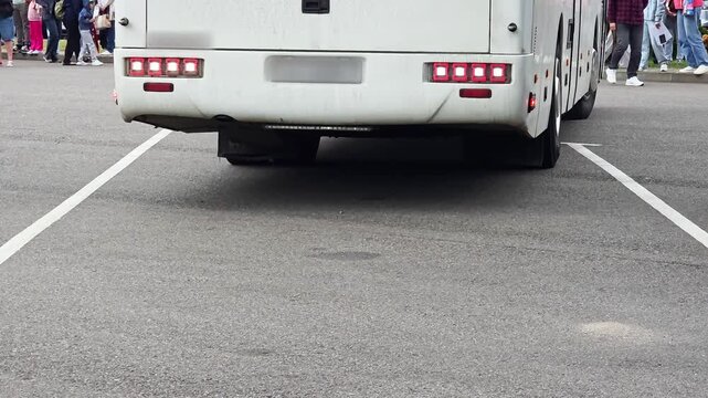 White bus rear view parked on asphalt parking lot with white parking lines, person walking past bus on left side, blurred people standing in background area, daytime scene, bus arrival at the station