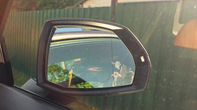 View from inside car showing side mirror reflection of rural road, green corrugated fence, lush green trees, and distant house under clear sky on sunny day