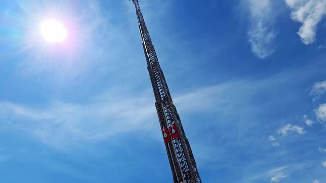 Extended aerial ladder of fire truck reaching high into bright blue sky with wispy clouds, powerful sun shining brightly overhead, rescue equipment.