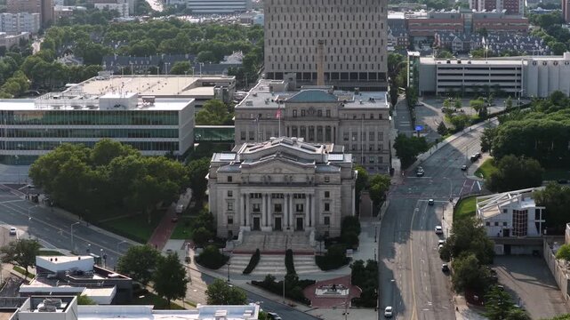 Domed courthouse with classical columns anchors downtown Newark plaza, glass office building and parking garage framing wide tree-lined boulevard with passing traffic. Aerial drone approaching shot