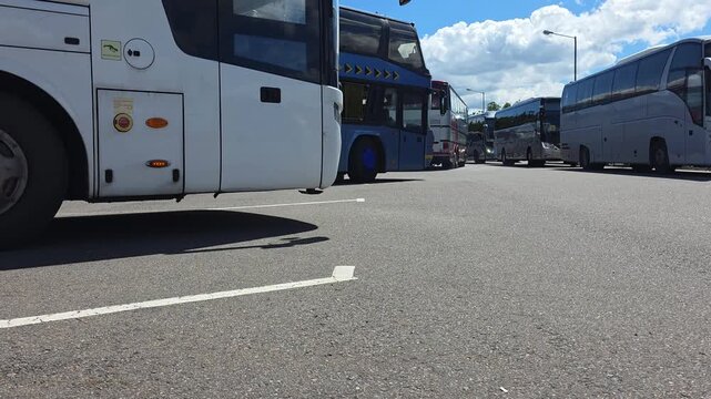 Multiple coaches and buses parked in rows on asphalt surface under blue sky with scattered clouds, showcasing transportation vehicles in large parking area on sunny day