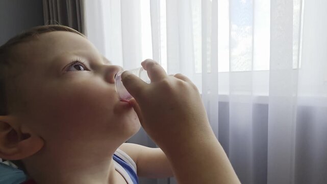 Young caucasian boy drinks cough medicine from a measuring cup, receiving medical treatment for illness, indoors by a bright window light, focusing on health and recovery.