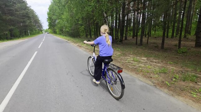 Young blonde girl with hair in ponytail rides bicycle on paved asphalt road during daytime, forest of tall green trees on right side of road, white lines marking lane