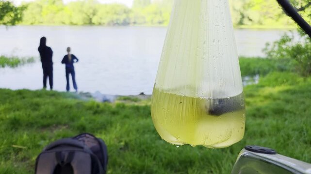 Live fish captured in clear plastic bag with water close up, in background two people stand by lake shore near small campfire, green grass and trees outdoor during day, outdoor recreation, copy space