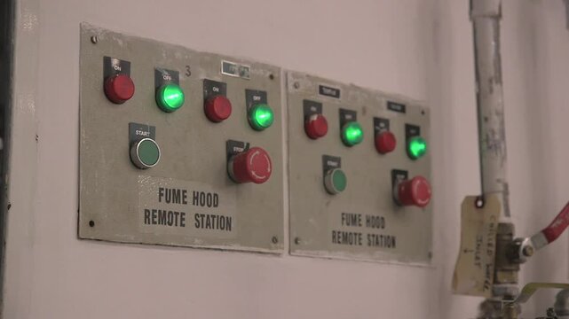 Fume hood remote station control panel inside a laboratory, with illuminated switches and emergency stop buttons on a wall-mounted unit.
