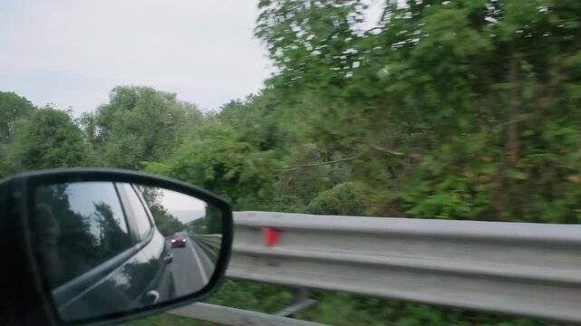 Slow motion side mirror view captures passing countryside and guardrail from moving vehicle. Blur of green forest landscape creates dynamic travel perspective..