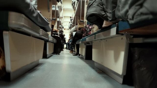 Interior view of Russian platzkart train car during night journey. Open-plan sleeper carriage. Long passage aisle with passengers occupying bunks and seats. Railway transport travel.