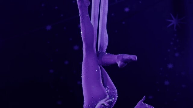 Female aerialist performing acrobatics on silk fabric, sparkling costume under purple light against starry night background, graceful movement of performer slow mo