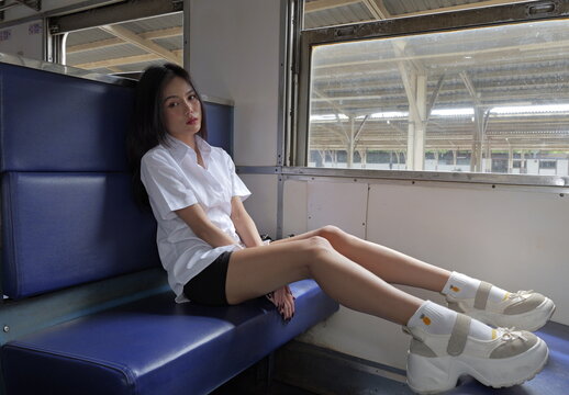 A young Asian woman wearing a university student uniform sits on a blue train bench seat with her legs extended across the bench