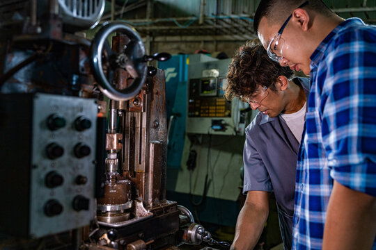 Thai father with curly hair and his teen son at the family machining workshop, dad bends low over the hobbing machine table, head down examining the setup, generational teamwork keeping