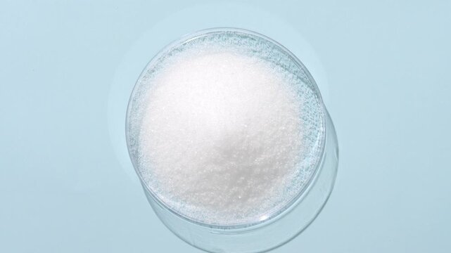 Top view capture of a mountain of white crystalline powder in a plastic petri dish on a light blue backdrop lit by moving light casting dynamic shadows | Magnesium oxide cosmetic ingredient concept