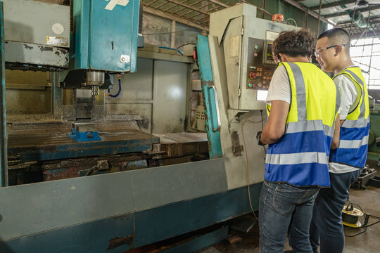 Middle aged Asian dad and teenage boy in a vintage family metal workshop, both stand back from the panel looking toward the milling table/spindle, dad's, father teaching son the trade