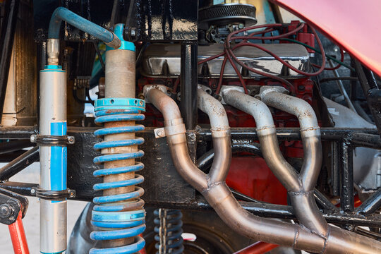 Close up shot of a custom off road race car engine compartment showing metal exhaust pipes, blue coilover shock absorber, and steel frame chassis. Mechanical detail of powerful racing vehicle parts.