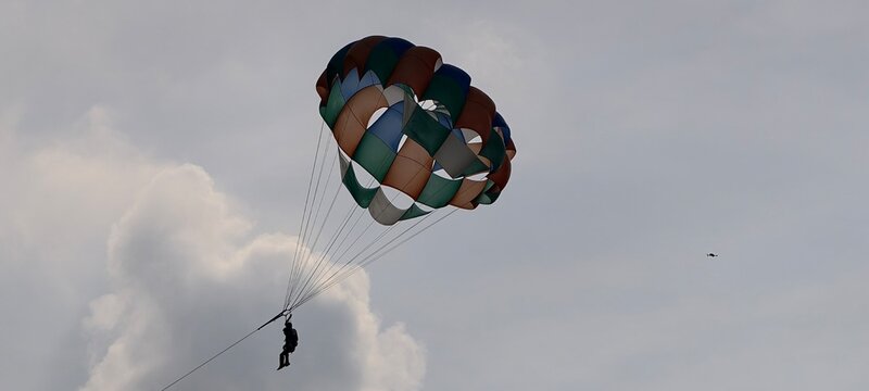 A person parasailing high in the sky with a multicolored parachute canopy in shades of green, blue, brown, and white. 