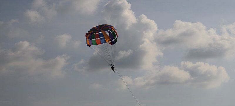 A vibrant parasail canopy in rainbow colors carries a person high above the ocean, suspended by cords and tow line against a partly cloudy sky. 