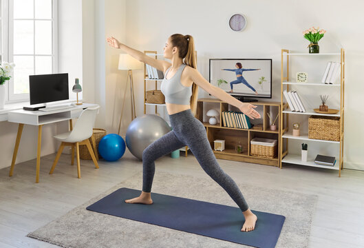 Woman practicing yoga workout at home television online. Full length female on a yoga mat mirrors a screen instructor for balance, flexibility and fitness. Concept shows mindful home exercise