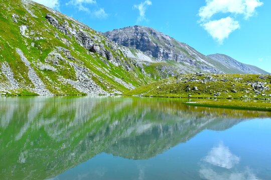 View of Oberere Lanischsee alpine lake with a reflection of the rock and green meadow covered mountains above in P&ouml;llatal, High Tauern, Carinthia, Austria