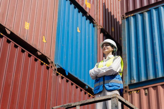 Female logistics engineer wearing hard hat standing with arms crossed in container shipping yard, Smiling woman inspector in safety vest looking at cargo containers in industrial port