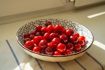 ripe red cherry in ceramic bowl on sunny windowsill at home. juicy cherry with water drops glowing in warm summer light. concept of cherry season and cozy childhood memories of tasty berry © RollAndSnap
