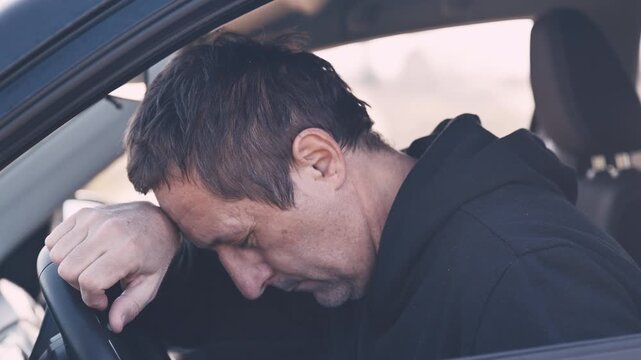Intimate portrait of a middle-aged man, seated in driver's seat of a car. Head resting heavily on forearm draped over steering wheel. 4K.