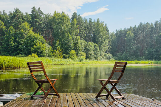 two wooden chairs on pier by calm lake for relaxation and forest view. cozy place for relaxation by water on sunny summer day. concept of relaxation and peaceful outdoor leisure in countryside nature