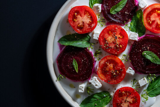 Gourmet salad with beetroot tomatoes and feta cheese. Top view closeup of colorful salad with sliced beetroot, fresh tomatoes, feta cheese, yogurt and mint leaves on white plate over dark background