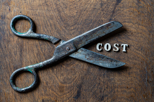 Vintage scissors cutting the word cost on wooden background, closeup. Top view conceptual shot of old rusty metal scissors cutting the word cost made of small letters on dark rustic wooden table