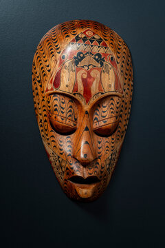 Handcrafted African style wooden mask on black background. Front view closeup of decorative hand carved wooden tribal mask with intricate black and red painted patterns hanging on dark textured wall