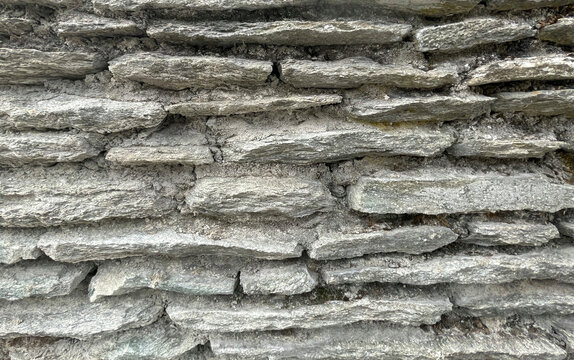 Detailed view of a textured stone wall construction with irregular shaped gray rocks and aged cement mortar binding them together, conveying a rustic and weathered facade, providing a sense of