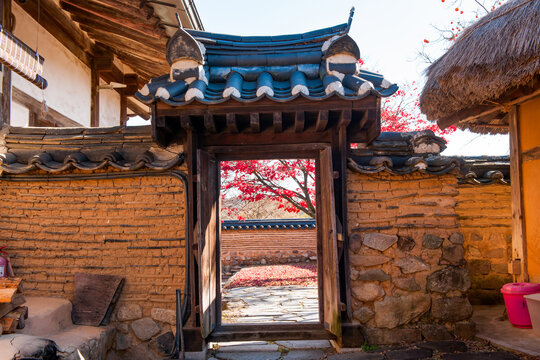 Traditional Korean hanok vernacular architecture horizontal composition framing vibrant autumn foliage through wooden Ilgakmun gate with stone-foundation mud wall