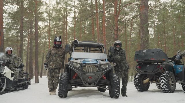 Three camo riders with atv and snowmobile in pine forest, backcountry guides prepping route and checking gear while standing beside buggy and sled, crisp snow under thick trunks, helmets and goggles,