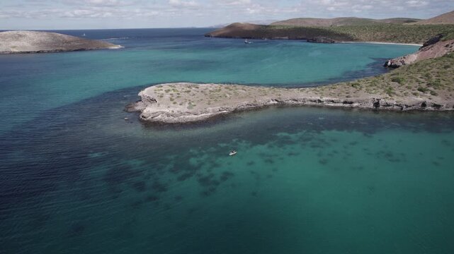 Stunning drone footage zooms in over turquoise waters and a rugged peninsula in Baja California Sur. Paddle boarders glide in the distance under bright, natural daylight, showcasing coastal serenity.