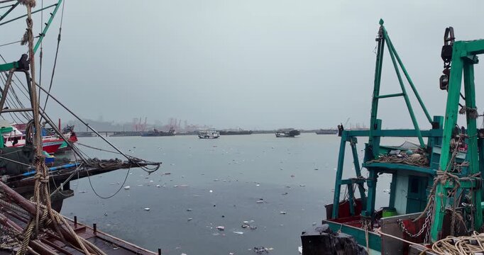 Wide view of the deck of a fishing boat cluttered with nets and ropes, overlooking a harbor polluted with floating plastic waste and debris during a rainfall, with ships visible in the misty distance