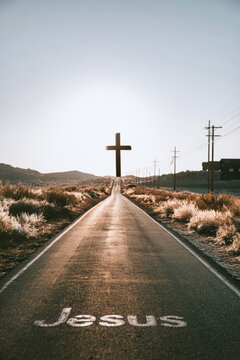 A straight road leads to a large cross at the end. The word Jesus is on the road with fields and distant hills surrounding the area