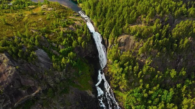 A breathtaking cinematic high-angle aerial tracking view capturing the powerful, twin cascading streams of L&aring;tefossen waterfall plunging through a dense, lush Norwegian forest.