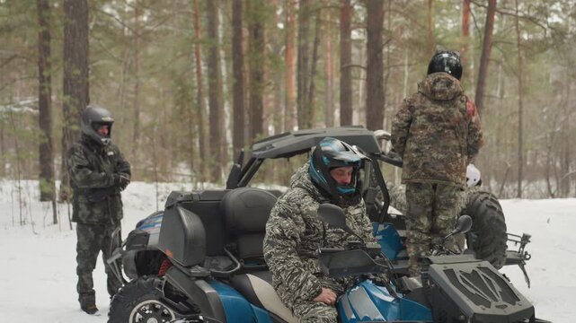Snowy forest atv riders gather around blue utv, helmeted crew prepare route, checking gear and throttle, camo jackets and goggles, pine trees and falling snow set brisk, tense expedition mood