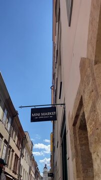 rectangular iron sign for mini market hangs from decorative bracket beside narrow lane, with plaster facades, shuttered windows, masonry details and blue sky above