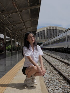 A young Asian woman wearing a university student uniform crouches on the edge of an open-air train platform