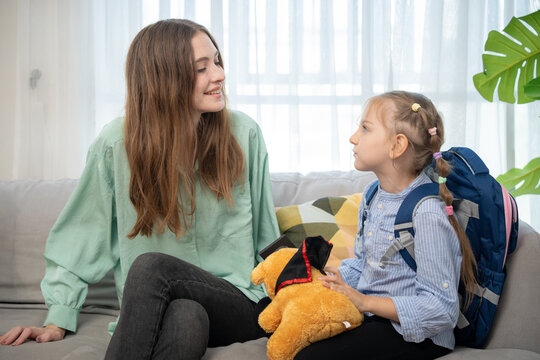 Loving mother looking at her little daughter and chatting warmly on sofa at home living room. Mother and schoolgirl daughter having sweet moment talking together on couch with teddy bear.