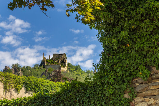 Blick auf die historische Burgruine D&uuml;rnstein in der Wachau gerahmt von efeubewachsenen Steinmauern und gr&uuml;nen Bl&auml;ttern unter blauem Sommerhimmel in &Ouml;sterreich.