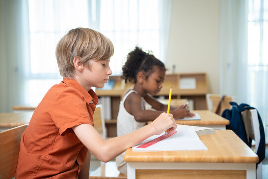 Young schoolboy using pencil and ruler to draw lines in notebook during lesson in elementary classroom. Focused student sitting at wooden desk drawing with ruler with classmate studying in background.