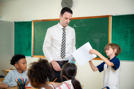 Confident young schoolboy standing up to hand his assignment paper to male teacher in classroom. Elementary student with glasses showing paper worksheet to teacher during group lesson.