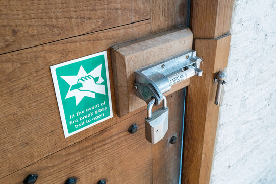 Detailed view of a security door, padlock and glass tubed security bolt seen within a famous British institution. The old wooden door leads to a car park and muster area for staff.