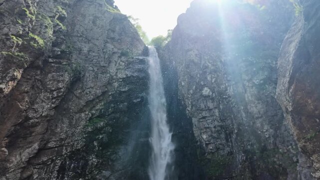 majestic cascading waterfall flowing down rough stone cliff faces with bright sunlight flare, mist and spray particles in the air, tranquil nature landscape for travel and meditation backgrounds