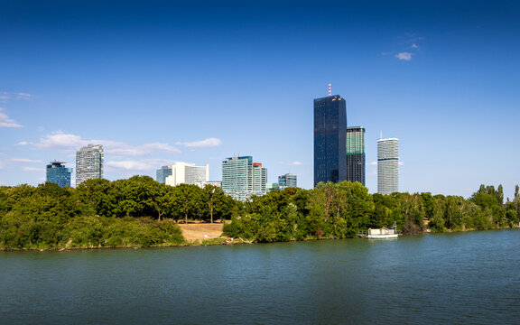 Die markante Skyline der Donau City in Wien mit dem DC Tower hinter dem gr&uuml;nen Ufer der Donauinsel bei strahlendem Sommerwetter und blauem Himmel.