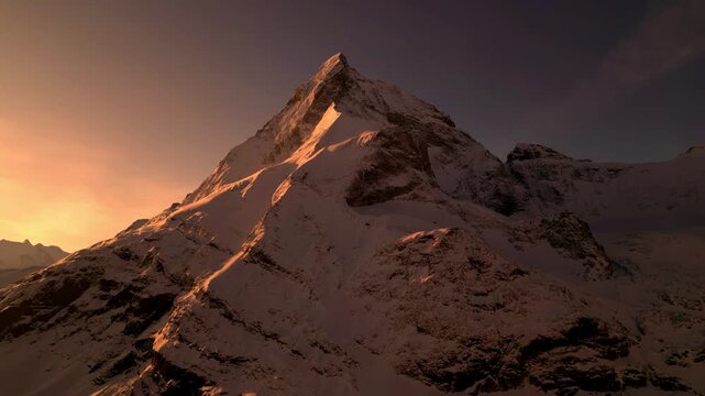 Aerial view of snow-covered mountain peak at golden hour with dramatic shadows and rugged alpine terrain in Zermatt, Wallis, Switzerland.