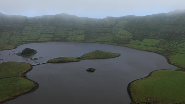 Aerial view of the Caldeirao crater lake with lush green volcanic slopes, stone walls, and small islets in Vila do Corvo, Acores, Portugal.