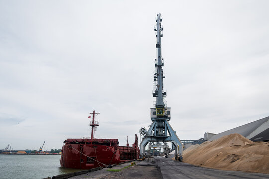 View of the Port of Ventspils featuring a large industrial crane and a red cargo ship docked at the pier with piles of sand nearby in Ventspils, Ventspils Municipality, Latvia.